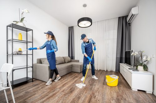 Professional cleaners in blue uniform washing floor and wiping dust from the furniture in the living room of the apartment. Cleaning service concept